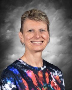 Smiling person with short, spiked hair wearing a colorful tie-dye shirt, against a gray background.