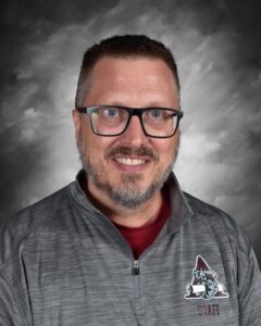 Smiling man with glasses and a beard, wearing a gray staff jacket, against a dark, cloudy background.