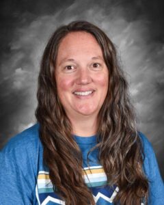 A smiling woman with long, wavy hair wearing a blue shirt with stripes, set against a gray background.