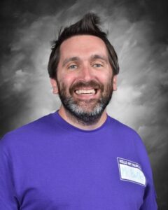 Smiling man with a beard, wearing a purple shirt and a name tag, set against a gray background.