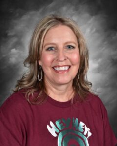 Smiling woman with long blonde hair wearing a maroon shirt with "HEY HEY!" text and dangling earrings.