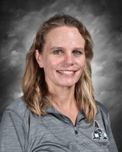 Headshot of a smiling woman with long blonde hair, wearing a gray athletic shirt with a logo. Background is gray.