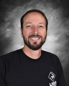 Smiling man with a beard, wearing a black t-shirt with a logo, against a gray background.