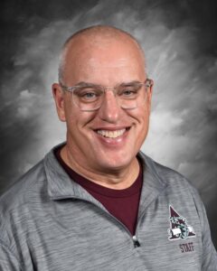 Smiling middle-aged man with glasses, wearing a gray staff jacket, set against a blurred gray background.