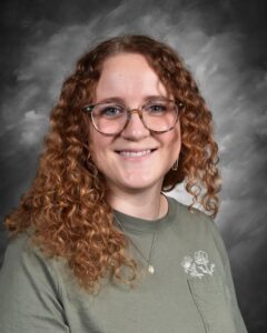 Smiling young woman with curly hair, wearing glasses and a green shirt, against a gray background.
