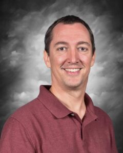 Smiling man with short dark hair wearing a maroon polo shirt, set against a gray, cloudy background.