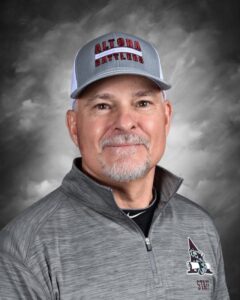 Portrait of a smiling man wearing a gray jacket and a cap with "Altona Battlers" logo against a cloudy background.