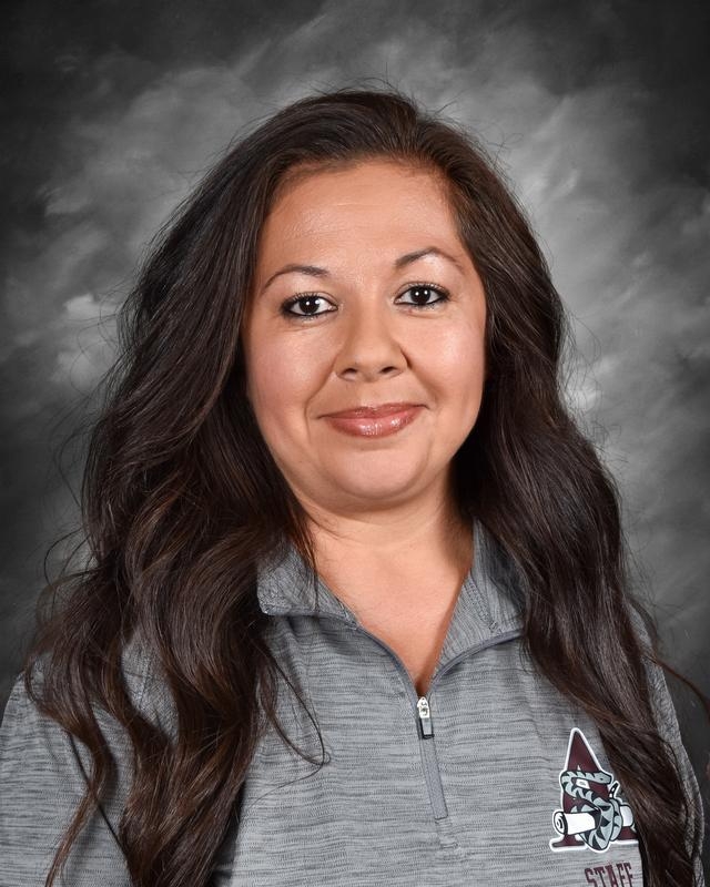 Close-up portrait of a woman with long dark hair, wearing a gray staff shirt with a logo, smiling against a gray background.