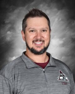 Smiling man with a beard, wearing a gray staff jacket, against a blurred gray background.