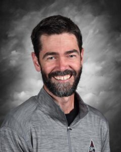 A smiling man with a beard, wearing a gray zip-up jacket, against a cloudy gray background.