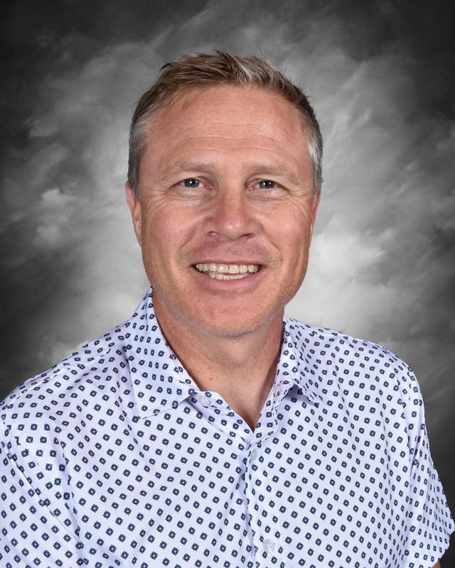 Smiling man with short hair wearing a patterned shirt, against a gray background.