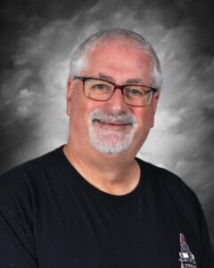 Smiling man with glasses and a beard, wearing a black shirt, against a gray background.
