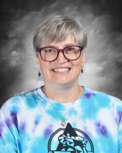 Mujer sonriente con cabello corto gris, gafas y camiseta teñida con la técnica tie-dye, sobre un fondo gris.