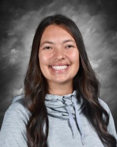 Smiling young woman with long, dark hair, wearing a light gray hoodie, against a soft gray background.