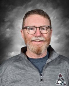 Smiling man with glasses and a beard, wearing a gray zip-up jacket, set against a cloudy background.