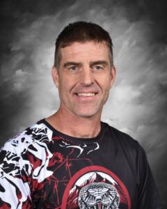 A man with short hair smiles, wearing a black and red graphic shirt with a snake design against a gray background.