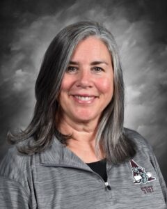 A smiling woman with long gray hair wears a gray jacket with a logo, set against a soft gray background.
