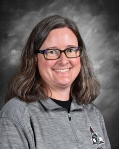 A smiling woman with long, wavy hair, wearing glasses and a gray athletic top, poses against a gray background.