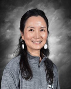 A woman with long black hair smiles, wearing a gray jacket and white earrings, against a dark, cloudy background.