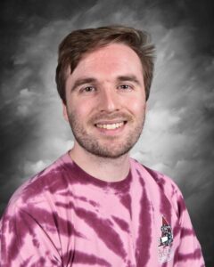 Smiling young man with short hair, wearing a pink tie-dye shirt, against a gray background.
