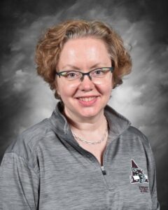 A woman with curly hair and glasses smiles, wearing a gray pullover with a staff logo against a dark background.