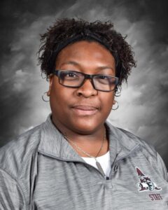 Portrait of a woman with curly hair wearing glasses and a gray staff shirt, set against a gray background.