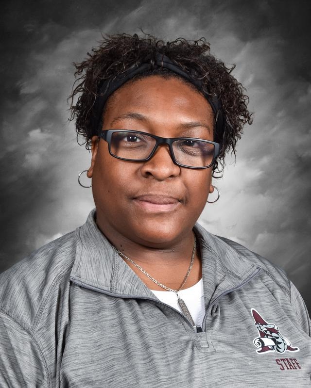 Portrait of a woman with curly hair wearing glasses and a gray staff shirt, set against a gray background.
