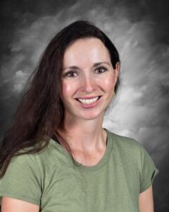 Smiling woman with long dark hair, wearing a green shirt, against a gray, cloudy background.