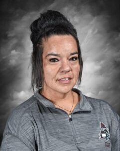 A woman with dark hair in a bun, wearing a gray zip-up shirt, smiles against a gray background.