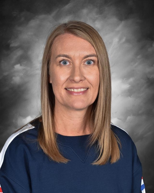 Portrait of a woman with long blonde hair, wearing a dark blue sweatshirt, smiling against a gray background.