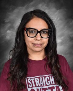 A woman with long, wavy hair, glasses, and a lip piercing, wearing a maroon shirt with white text.