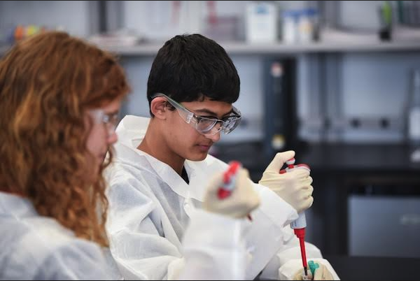 Two students in lab coats and goggles conduct an experiment in a laboratory, using pipettes to measure liquids.