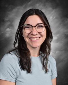 Smiling woman with long dark hair and glasses, wearing a light blue shirt, against a gray background.