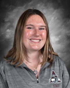 Smiling young woman with shoulder-length hair, wearing a gray staff shirt, against a dark background.