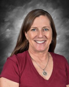 A smiling woman with long brown hair wears a maroon shirt and a necklace, set against a gray background.