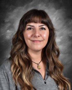 Portrait of a woman with long, wavy hair, wearing a gray shirt and a necklace, smiling against a gray backdrop.