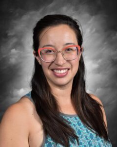 Mujer sonriente con cabello largo y oscuro, gafas con montura rosa y blusa azul estampada sobre un fondo gris.