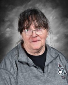 Older woman with glasses, wearing a gray zip-up top, smiling against a gray background.