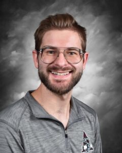 Smiling man with glasses and a beard, wearing a gray athletic shirt with a logo, against a gray background.