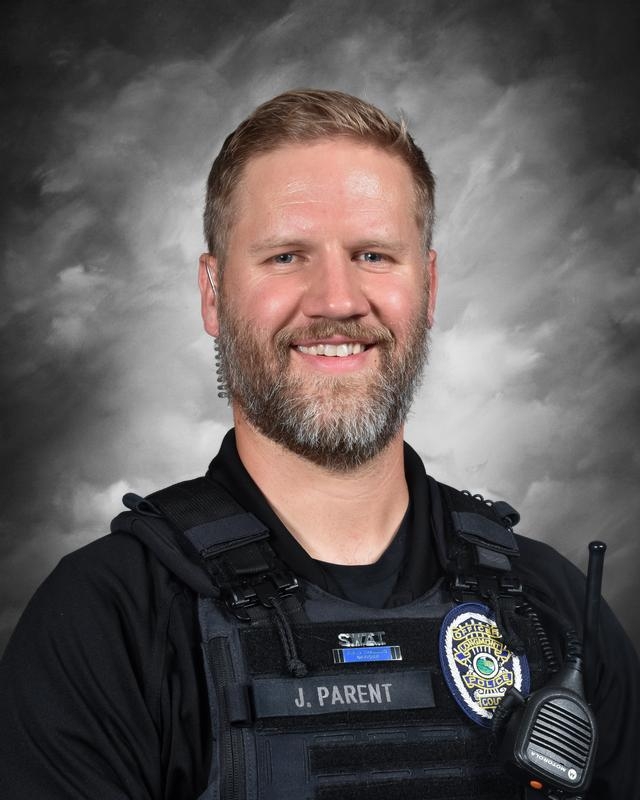 Portrait of a smiling police officer in tactical gear, with a radio and badge, against a gray background.