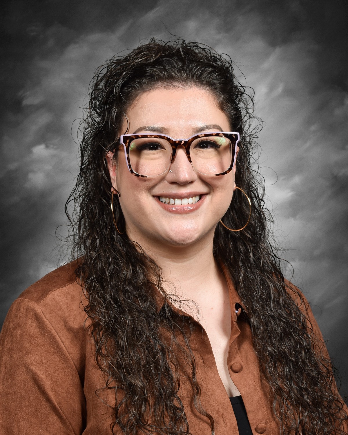 A smiling woman with curly hair wears large glasses and hoop earrings, dressed in a brown jacket against a gray backdrop.