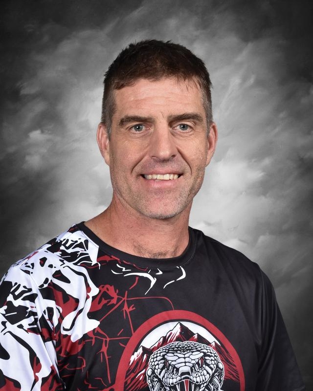 Un hombre con el pelo corto y una sonrisa amable, vestido con una camiseta estampada en negro y rojo, sobre un fondo gris.