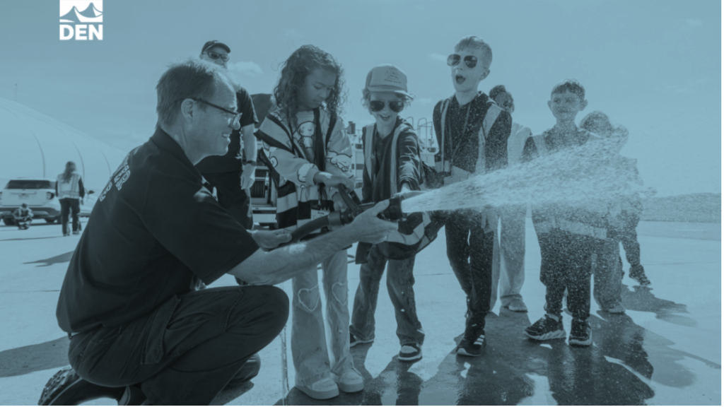 A firefighter demonstrates a hose spray to a group of excited children at an outdoor event.