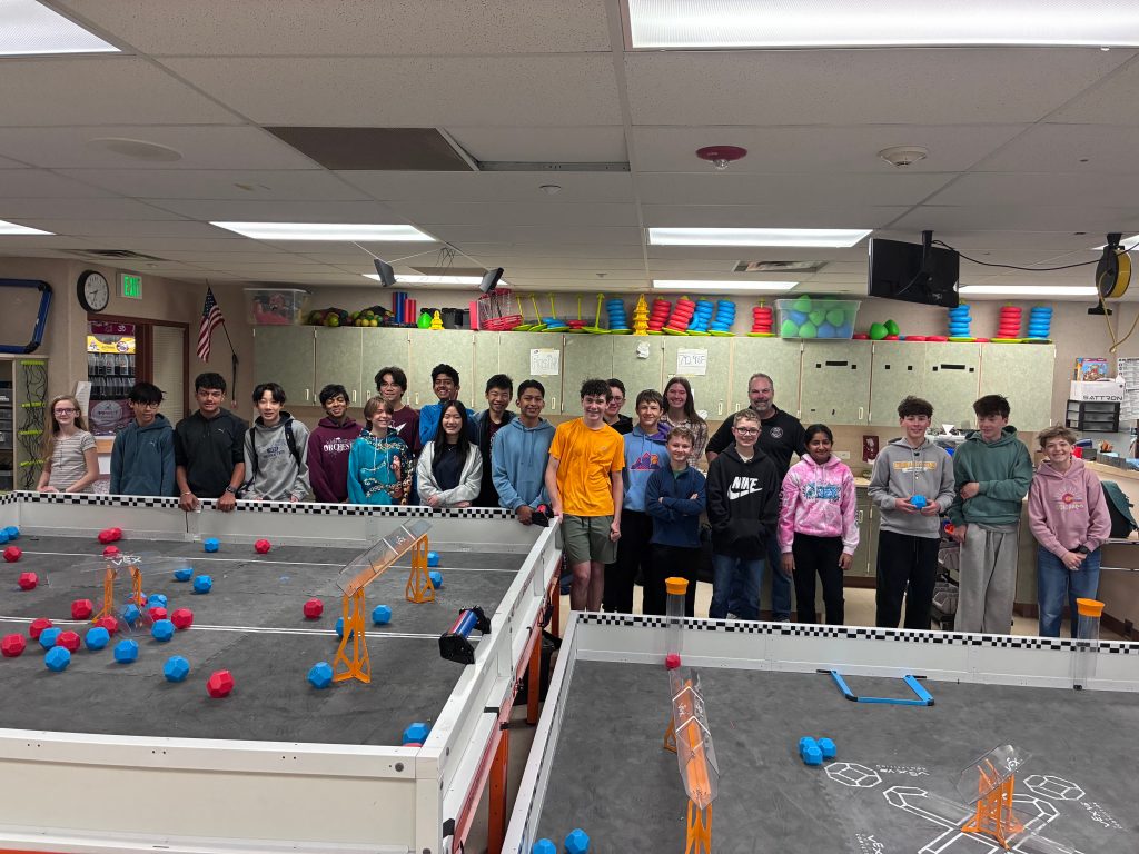 A group of students and adults stands in a classroom with a robotics competition setup featuring colorful game pieces.