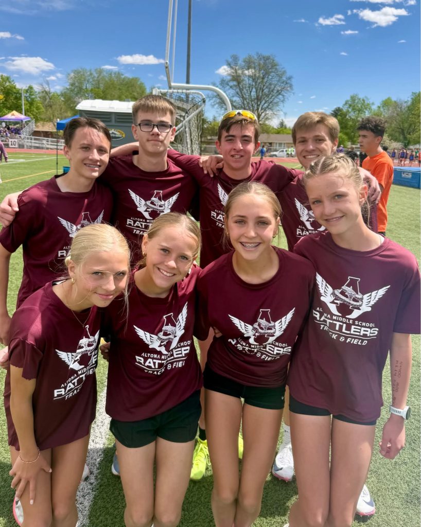 A group of eight middle school students in maroon track shirts pose together on a sunny field, smiling happily.