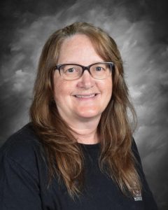A smiling woman with glasses, long brown hair, and a black shirt against a gray background.