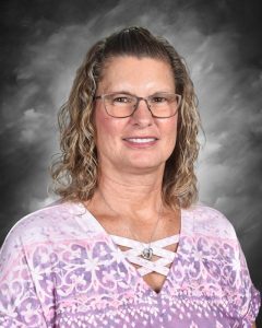 Person with curly hair and glasses, wearing a pink patterned top, smiles against a gray background.