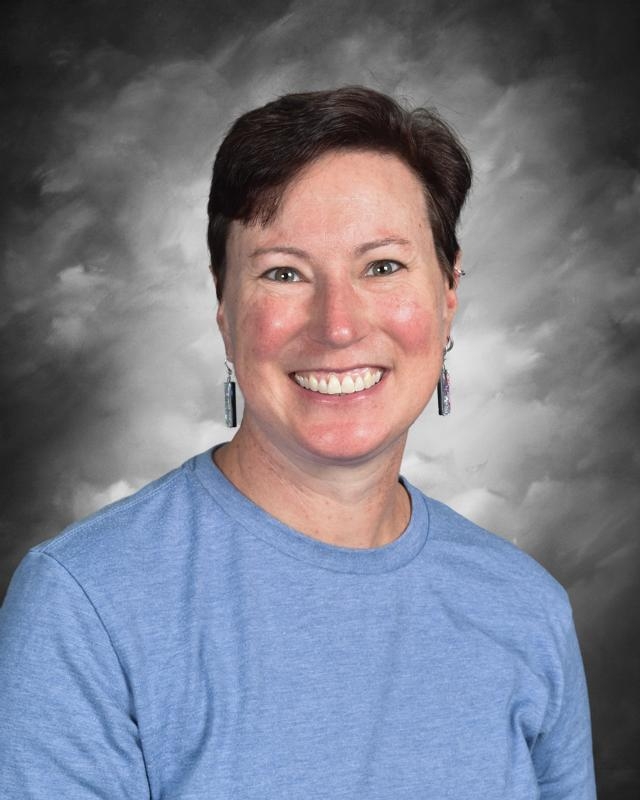 Person smiling, wearing a blue shirt and earrings, with a cloudy gray background.